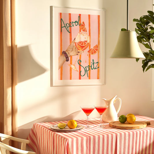 Framed artwork with 'Aperol Spritz' design above a table set with drinks and fruit.