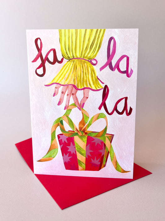 A greeting card with a whimsical illustration of a yellow curtain, pink hands, and a red gift box with green ribbon, placed on top of a red envelope against a neutral light gray background.