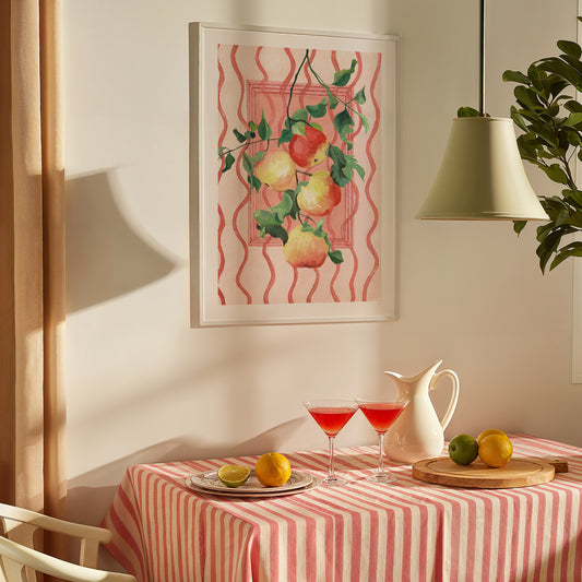Dining table with a striped tablecloth, pitcher, glasses, and fruit, with a framed artwork of apples on the wall.