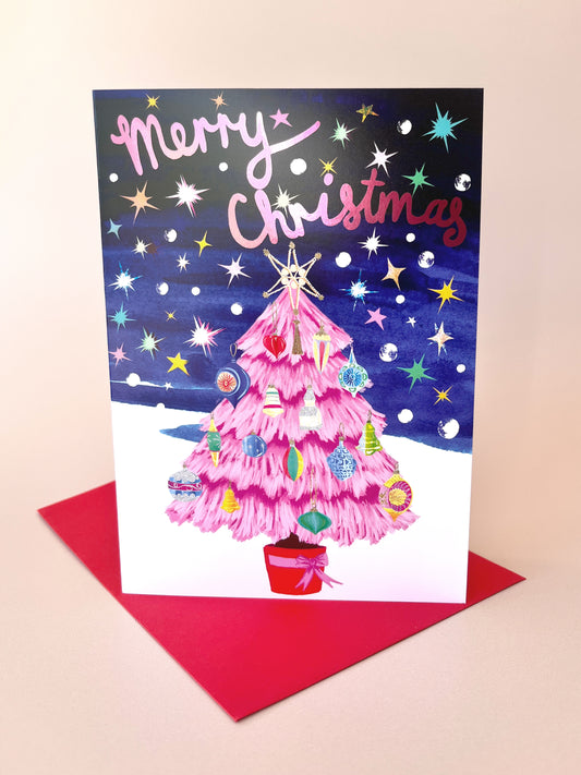 A Christmas greeting card with a pink tree illustration and the words 'Merry Christmas' on a dark starry background, displayed upright on a red envelope against a light beige backdrop.