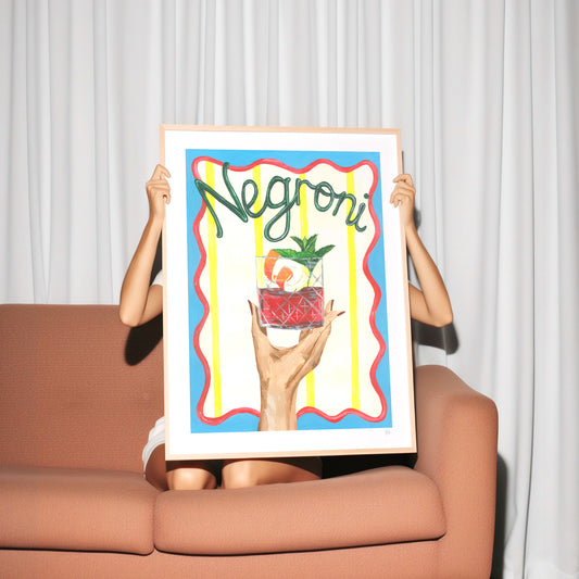 Person holding a colorful poster with 'Negroni' text and cocktail illustration in front of a white curtain.