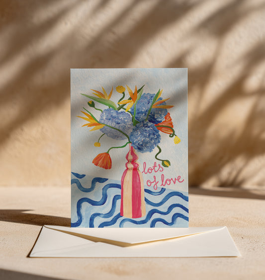 A square greeting card with a colorful floral illustration in a pink vase and the words 'lots of love,' displayed upright on a beige surface with an envelope in front. The scene is lit by warm natural light with soft shadows.