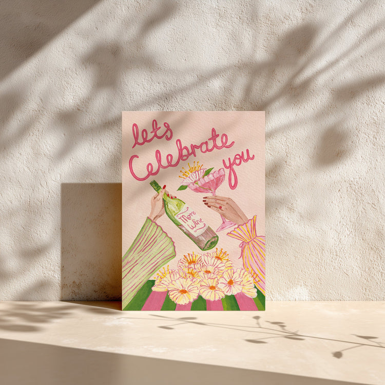 A vertical greeting card with the text 'let's Celebrate you' in pink script, featuring an illustration of hands holding a bottle and a glass of wine, surrounded by flowers, displayed on a beige surface with textured plaster walls and leaf shadows.