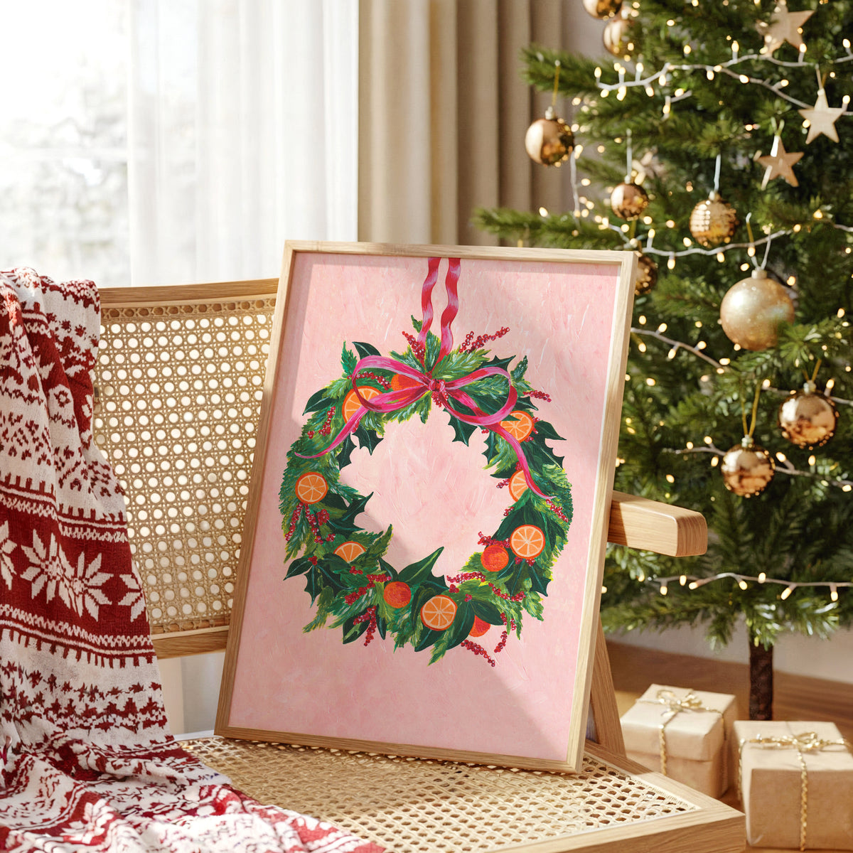 A framed Christmas wreath art print with a pink background, green holly and ivy, orange citrus slices, and a pink bow, leaning on a woven chair in a decorated holiday room next to a Christmas tree and wrapped gifts.