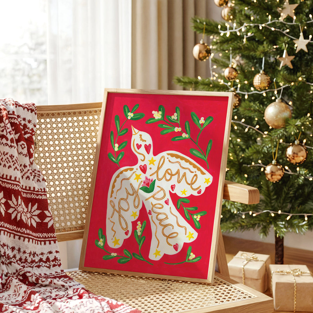 A framed Christmas art print featuring a white dove with 'Love & Peace' written on it, surrounded by green holly leaves and red background, styled on a cane chair next to a decorated Christmas tree.