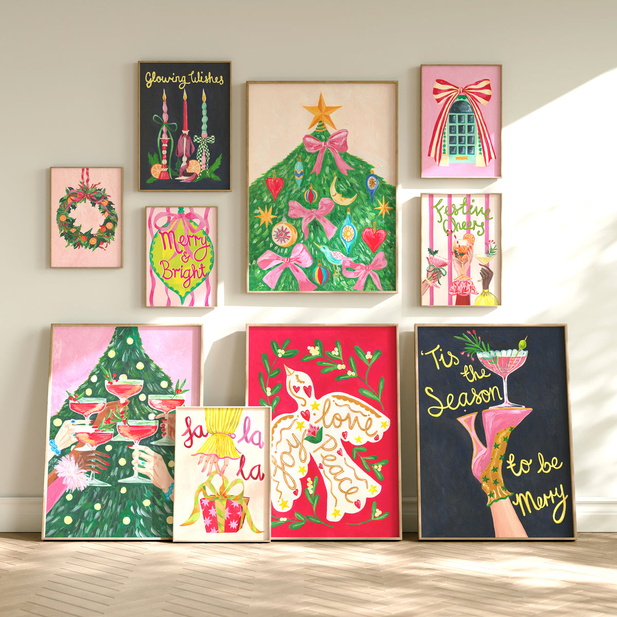 A collection of colorful framed art prints arranged on a light beige wall above a light wood floor. The prints feature whimsical illustrations including a green Christmas tree with pink bows, a wreath, cocktails, gifts, and playful text such as 'My Brain', 'Life is Cool', and 'Fun Season to be Merry'.