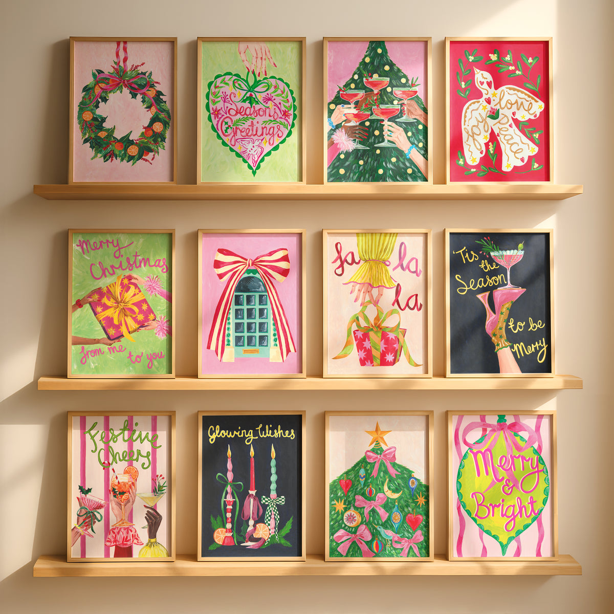 Twelve framed Christmas art prints are arranged on three light wood shelves against a warm beige wall. Each print features a different festive illustration, including a wreath, a heart-shaped Christmas tree, a Christmas tree with champagne glasses, a dove, a gift box, a door with a red bow, 'fa la la' text, a cocktail glass with 'It's a Season to be Merry', a cocktail glass with 'Fesrtive Cheers', glowing candles, a decorated Christmas tree, and a bright green bauble with 'Merry & Bright'.