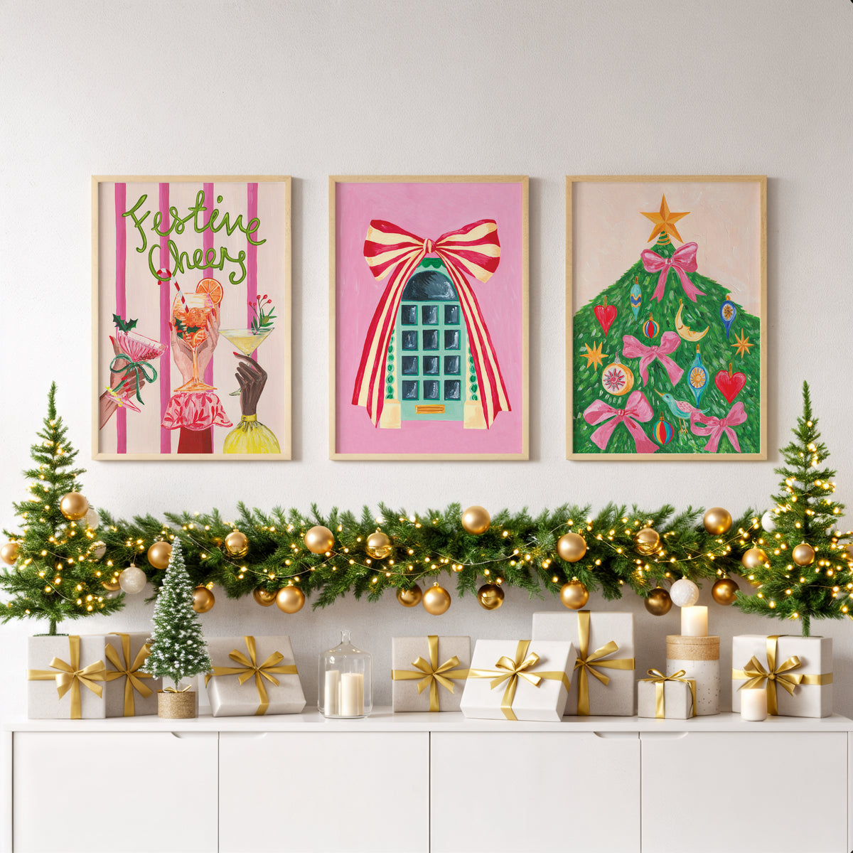 Three framed Christmas art prints are displayed on a white wall above a decorated holiday mantel with garlands, gold ornaments, wrapped gifts, and candles.