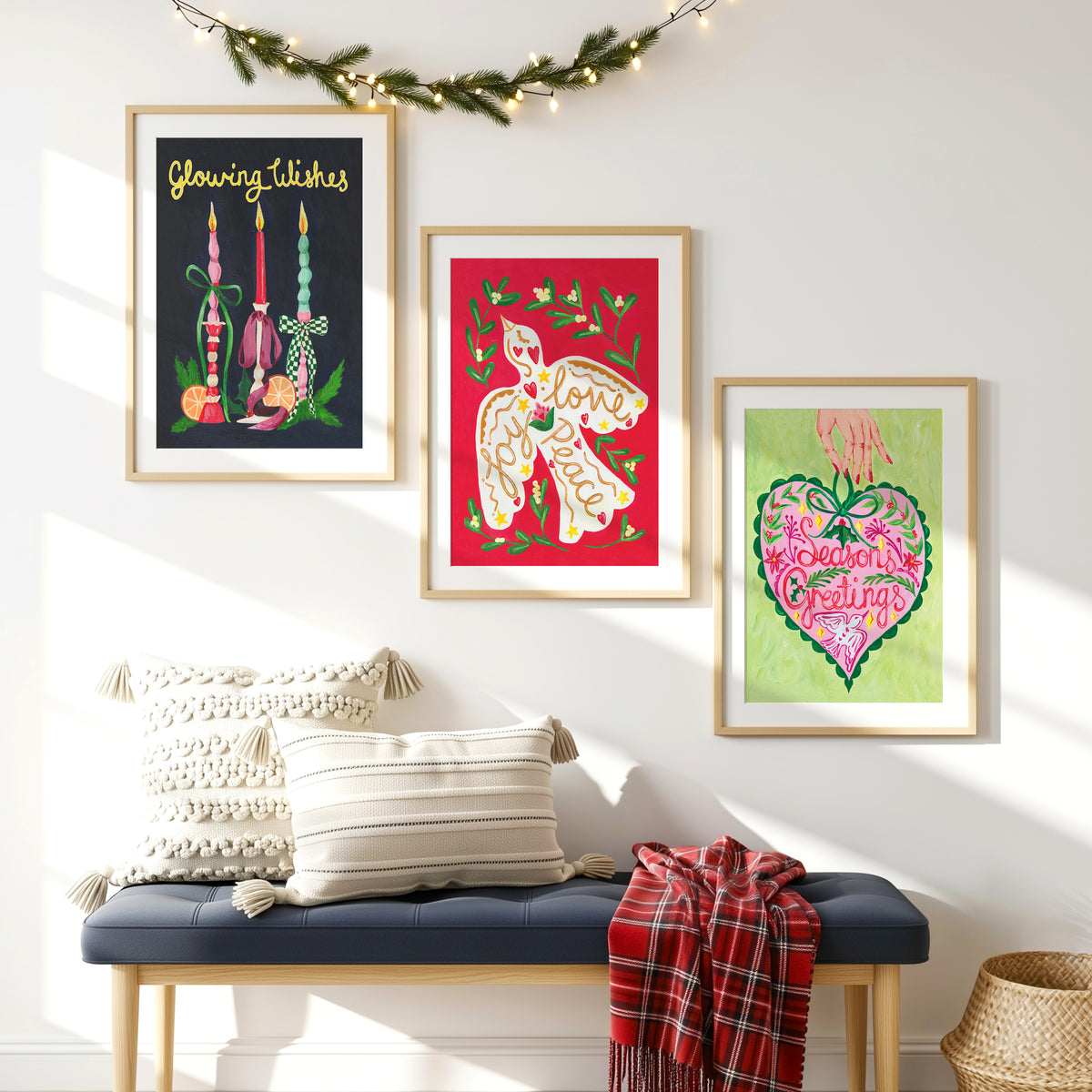Three framed art prints with Christmas-themed designs are displayed on a white wall above a blue bench with pillows and a red plaid throw. A green garland with string lights hangs across the top.