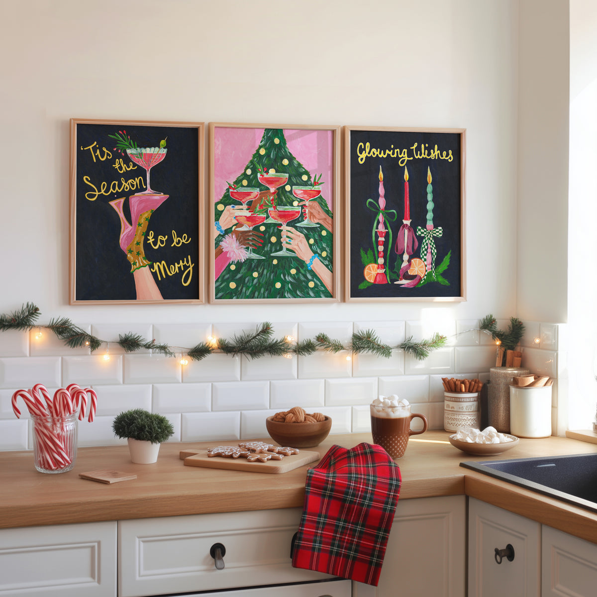 Three framed Christmas-themed art prints hang on a white subway tile kitchen wall above a wooden counter decorated with holiday items, including candy canes, a tart, and a red plaid towel.