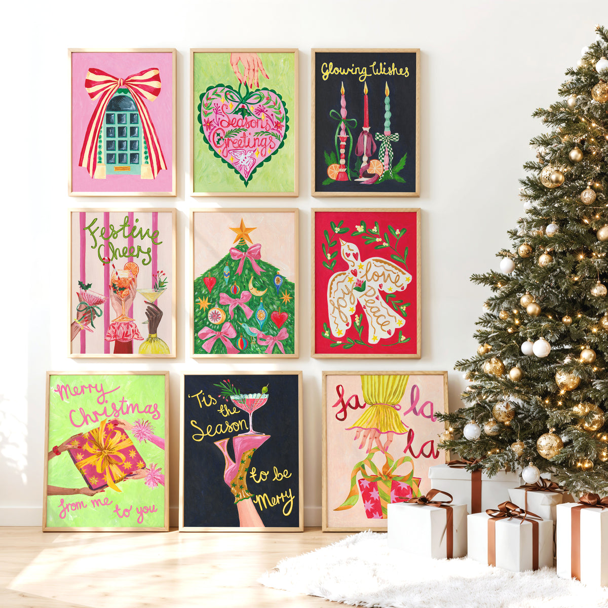 Nine framed Christmas art prints arranged in a 3x3 grid on a white wall, with a decorated Christmas tree and wrapped gifts to the right. The prints feature colorful festive illustrations including a bow, a heart, candles, a bird, a Christmas tree, gifts, and holiday phrases.