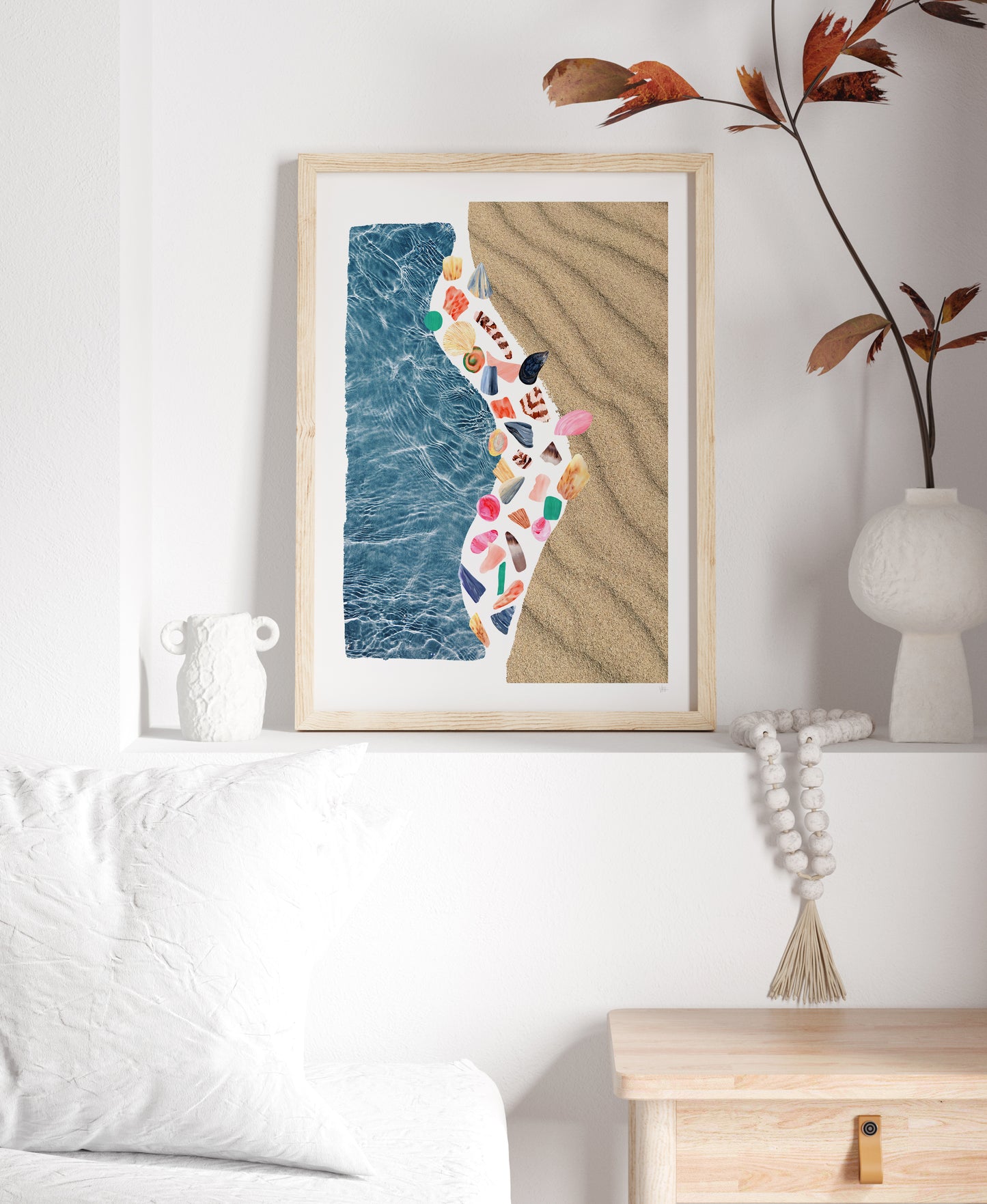 Framed artwork of a beach scene with blue water, sand, and colorful shells on a white wall.