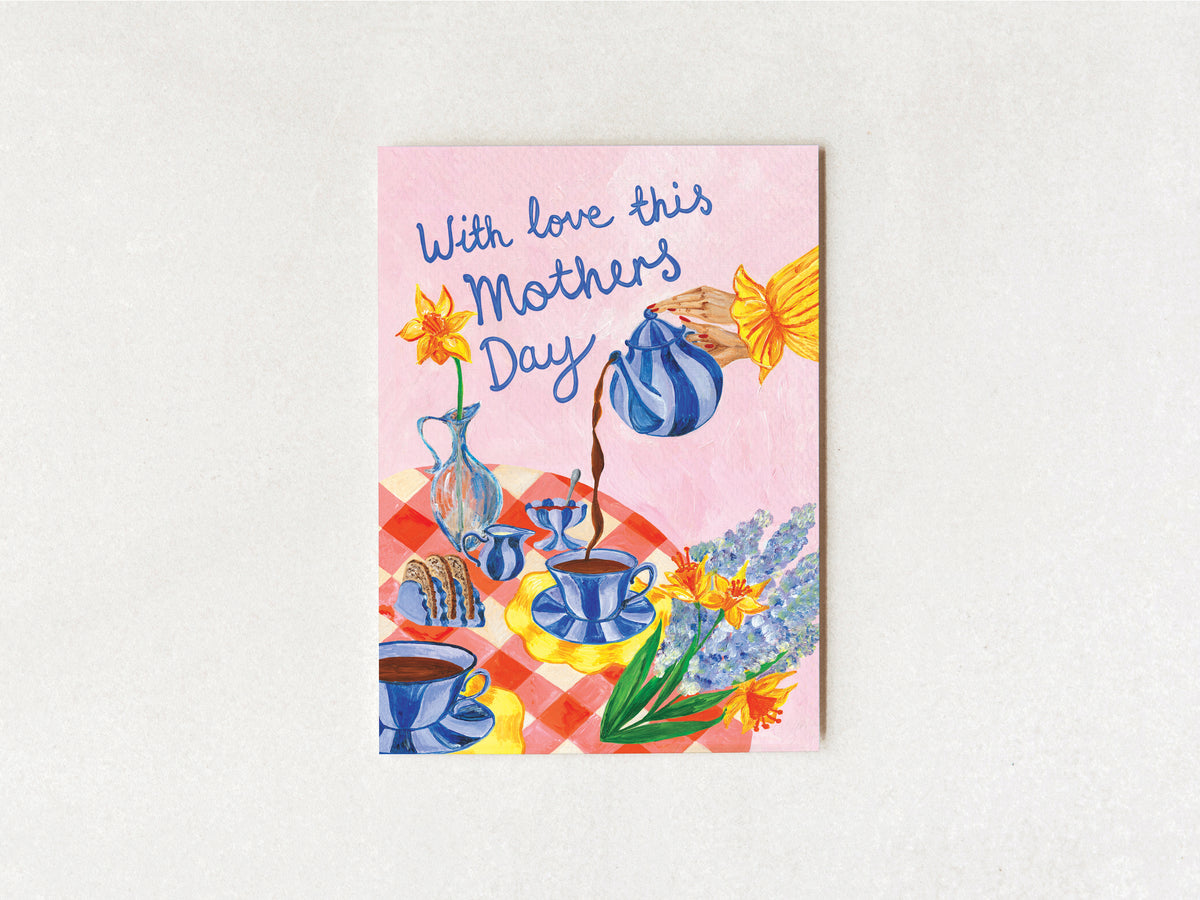 A square greeting card with a pink background and an illustration of a tea-time scene, reading 'With love this Mothers Day,' displayed against a light textured wall.