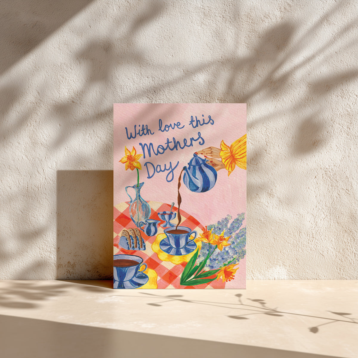 A square greeting card with a pink background and a colorful illustration of teacups, flowers, and the text 'With love this Mothers Day' stands against a textured beige wall with leaf shadows.