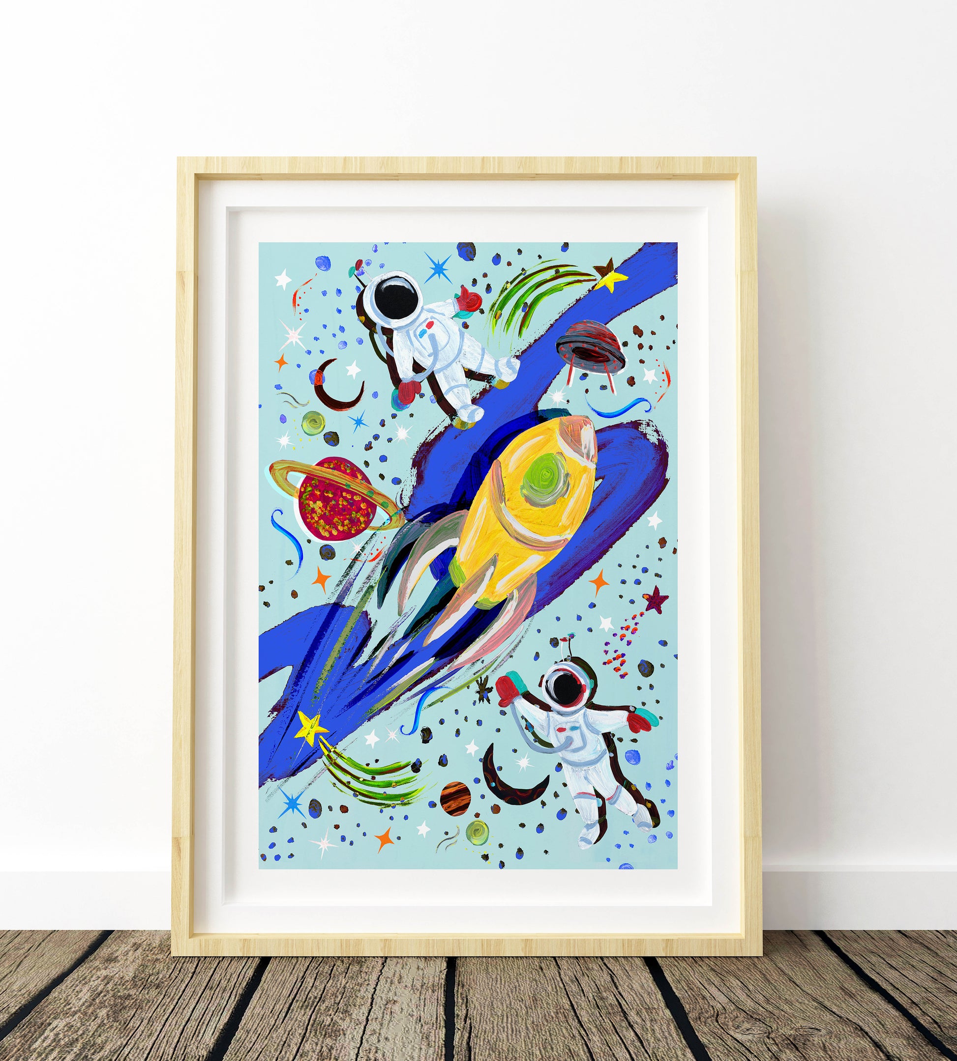 Framed artwork of a colorful space-themed painting with astronauts and rockets on a light blue background.