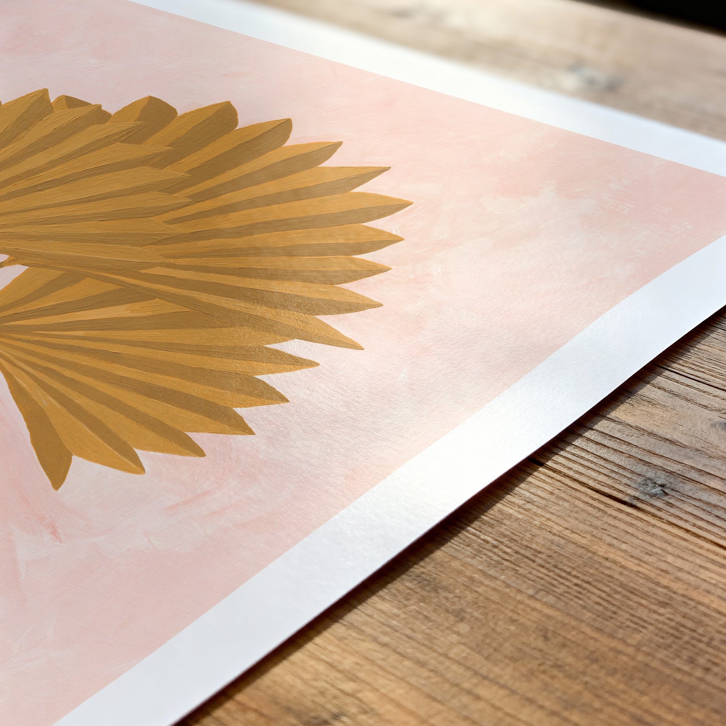 Gold palm leaf design on a pink background with a white border, placed on a wooden surface.