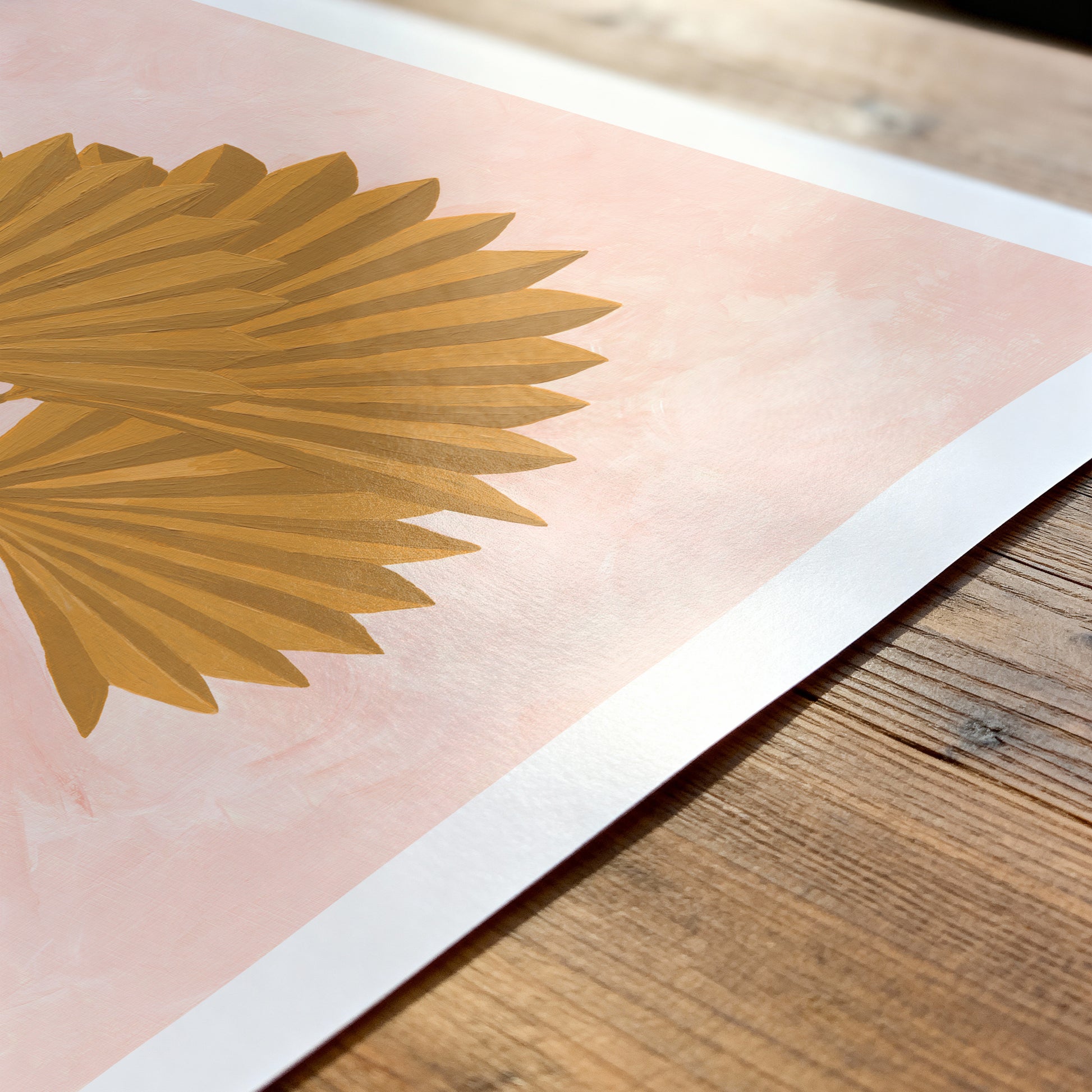 Gold palm leaf design on a pink background with a white border, placed on a wooden surface.