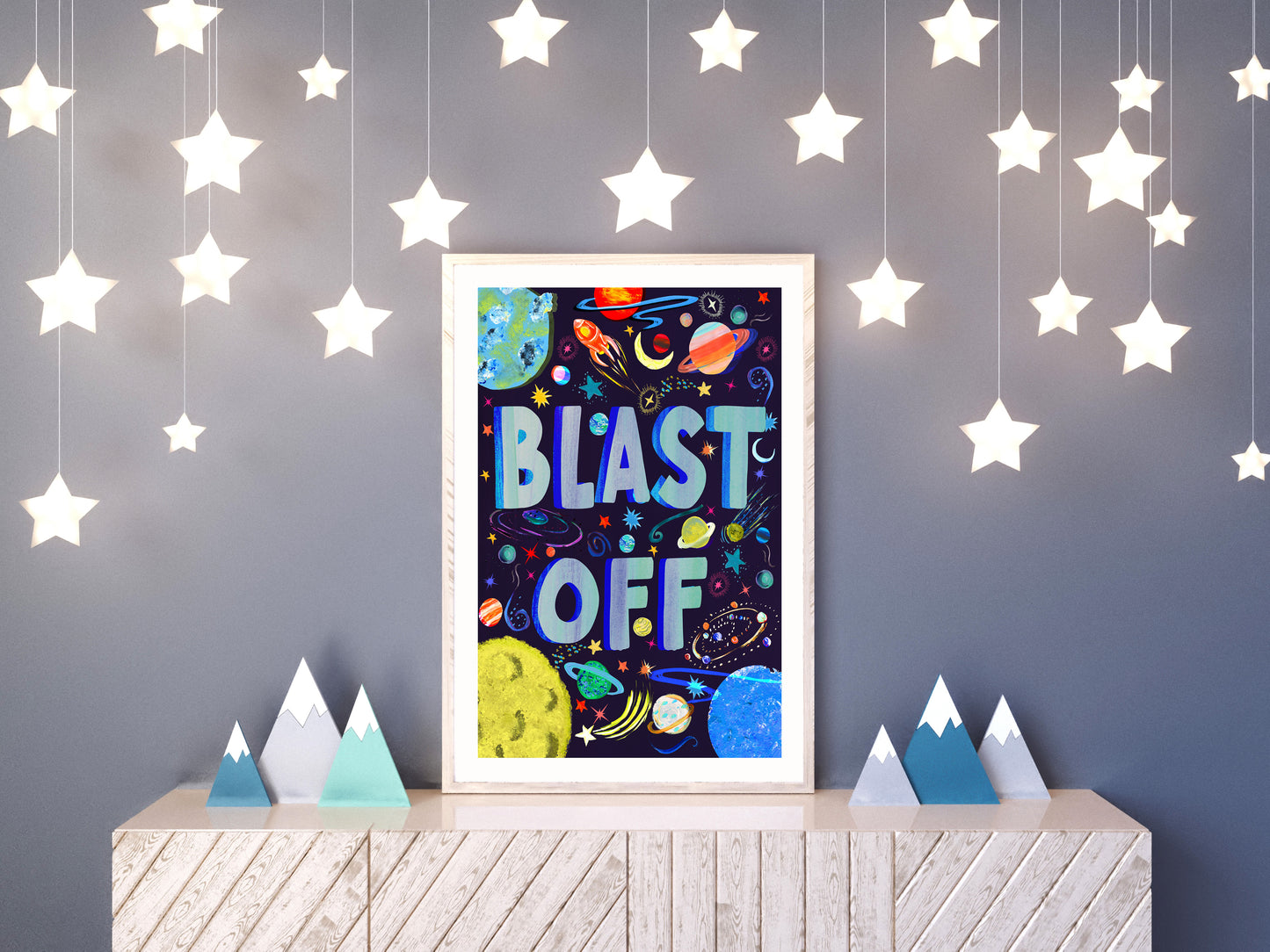 Framed space-themed poster with 'BLAST OFF' text on a wall with hanging star lights.