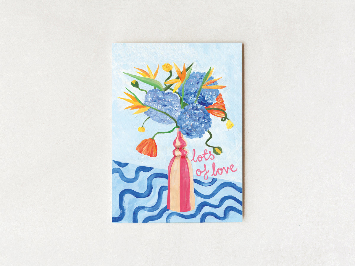 A vertical greeting card featuring a colorful illustration of a flower bouquet in a pink vase against a light blue background, with the words 'lots of love' in pink script. The card is displayed flat against a textured off-white background.