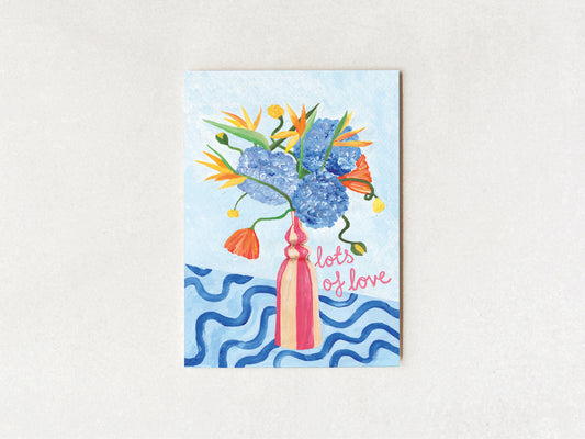 A vertical greeting card featuring a colorful illustration of a flower bouquet in a pink vase against a light blue background, with the words 'lots of love' in pink script. The card is displayed flat against a textured off-white background.