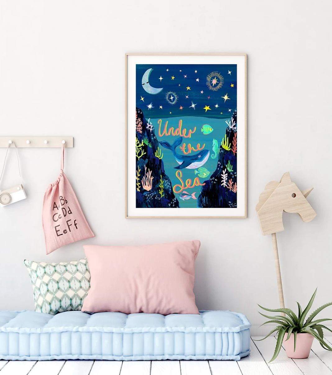 Framed artwork titled 'Under the Sea' in a room with a light blue bench, pink pillow, and wooden unicorn decoration.