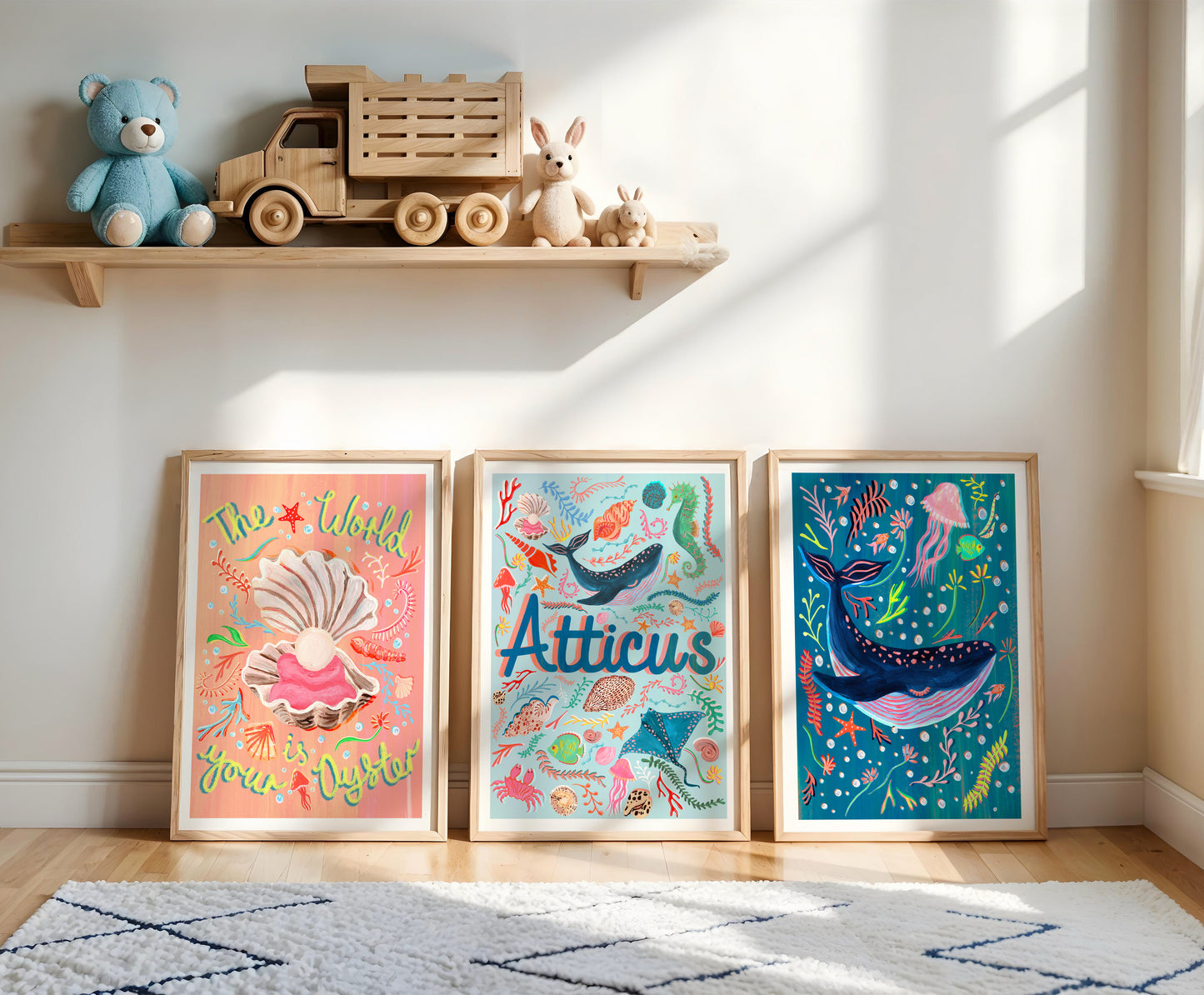 Three framed ocean themed prints 'the world is your Oyster' shell illustration, name atwork with sealife illustrations and a blue whale on a wooden floor with toys on a shelf in the background.