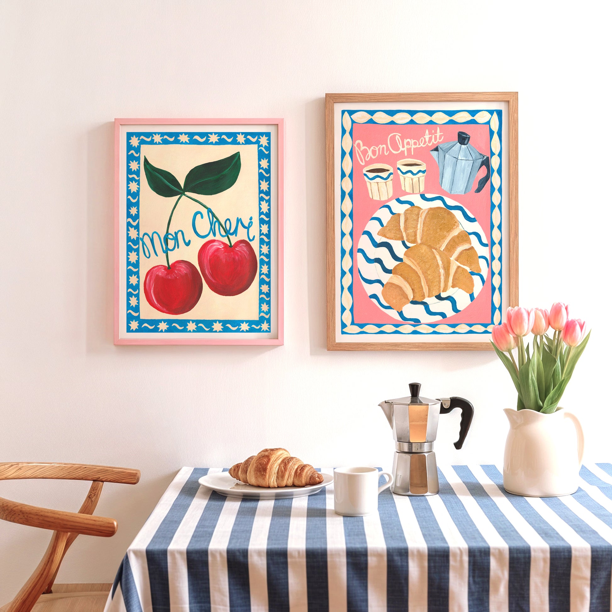 Two framed colourful kitchen artworks on a wall above a table with a striped tablecloth, coffee maker, and flowers.