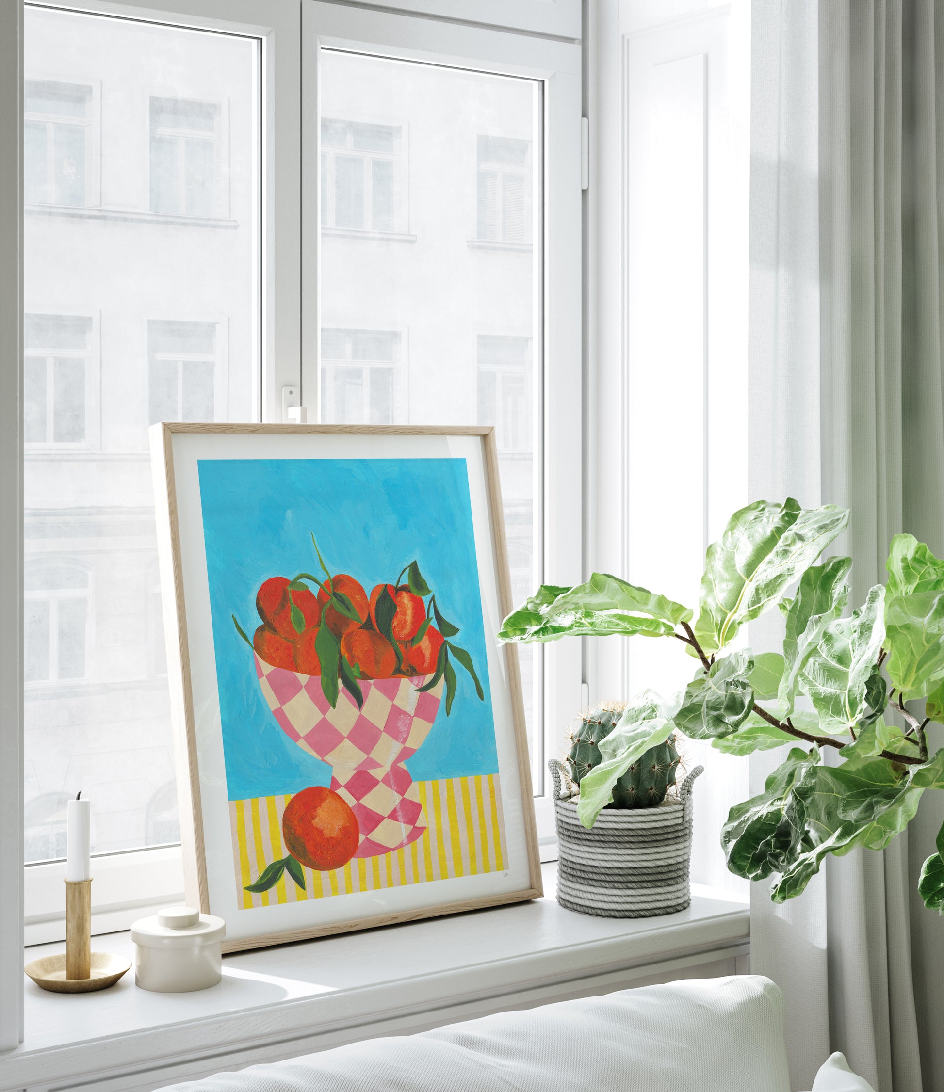 Framed artwork of oranges in a checkered bowl on a windowsill with a plant and candles.