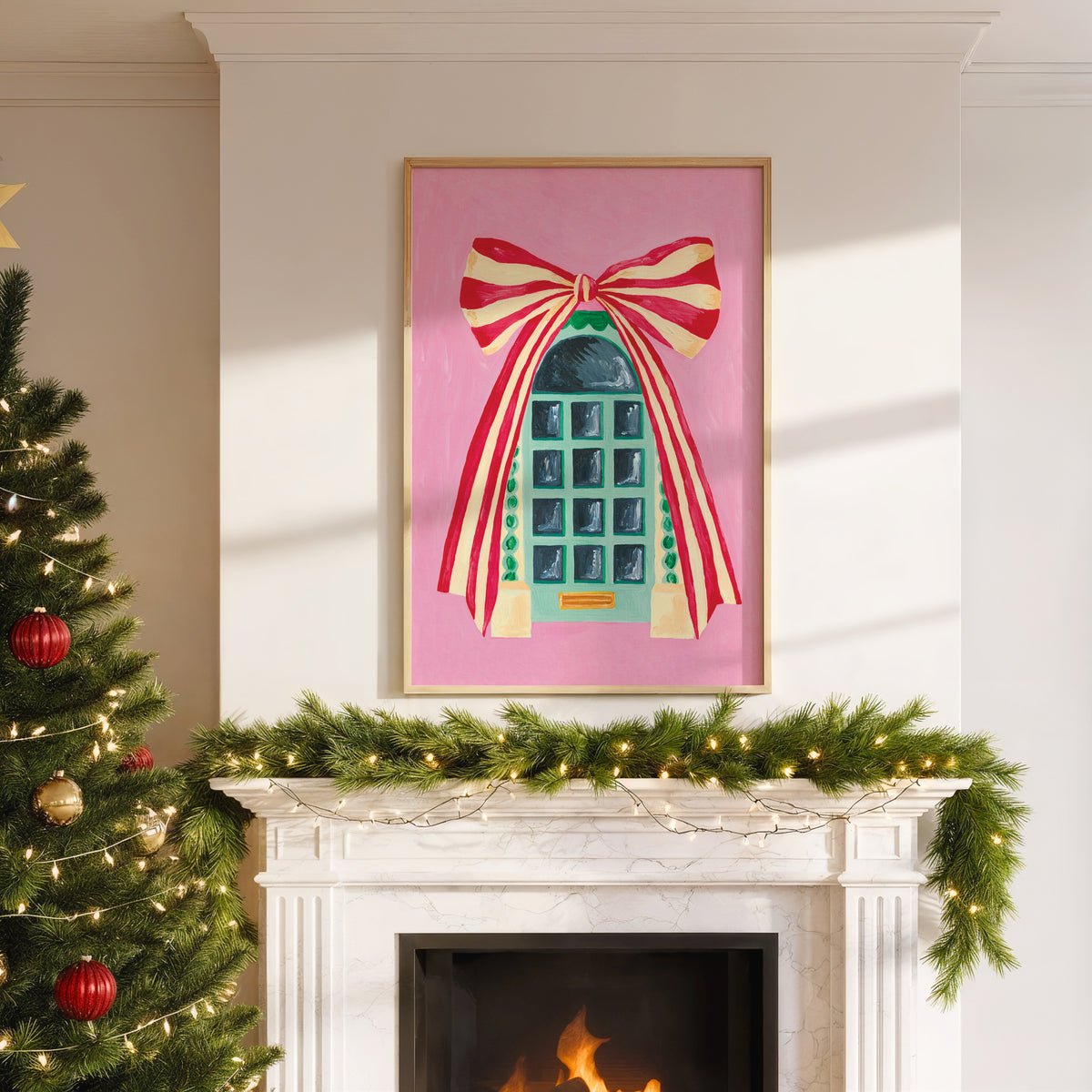 A framed art print featuring a stylized teal telephone booth under a large red and cream striped bow on a pink background, displayed on a white fireplace mantel above a lit fire, with a decorated Christmas tree to the left.
