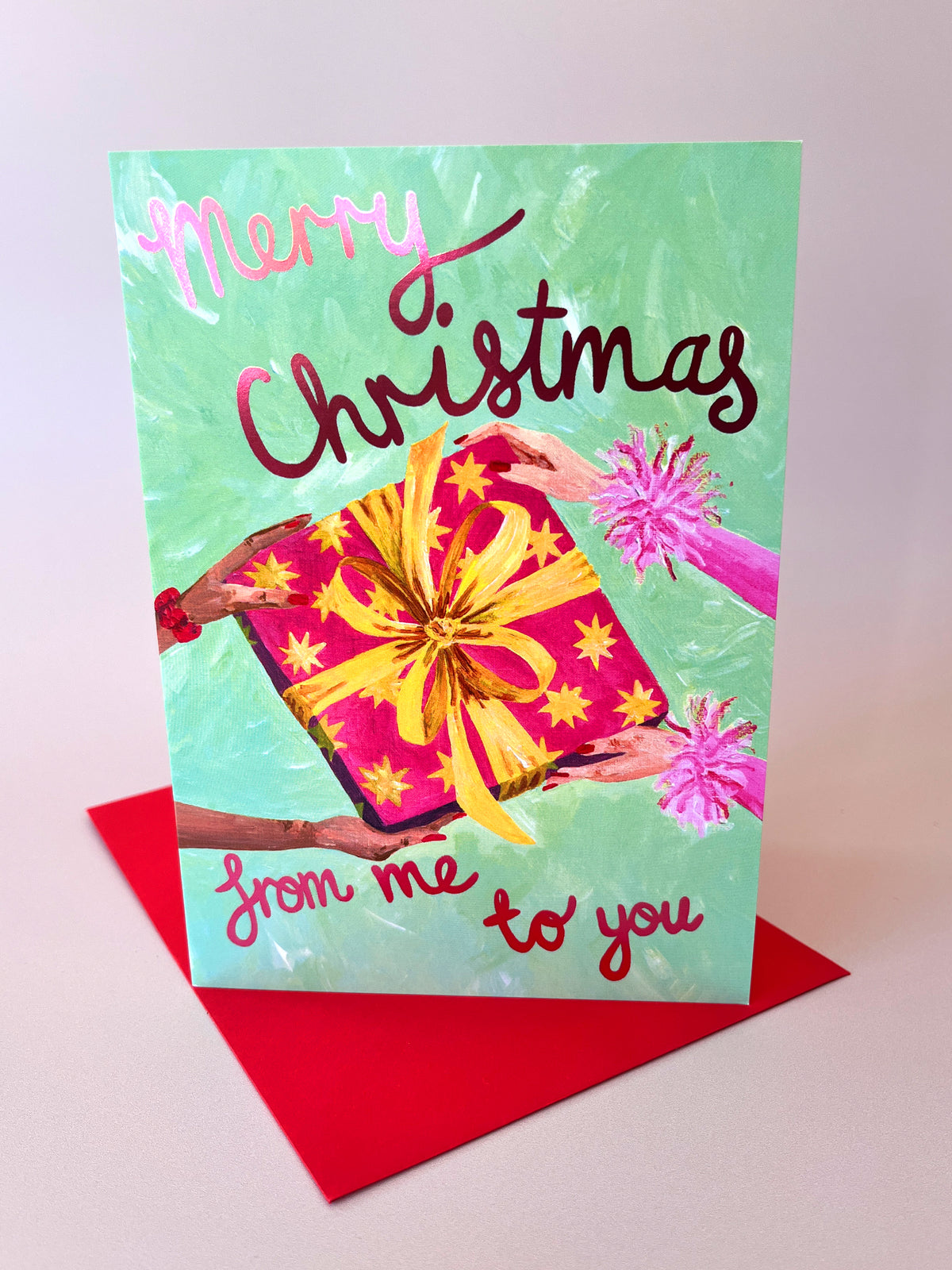 A Christmas greeting card with a mint green background, featuring the text 'Merry Christmas from me to you' in pink and burgundy script. The card is illustrated with hands holding a red gift box with a yellow bow and stars, and is shown standing upright on a red envelope against a light pink background.