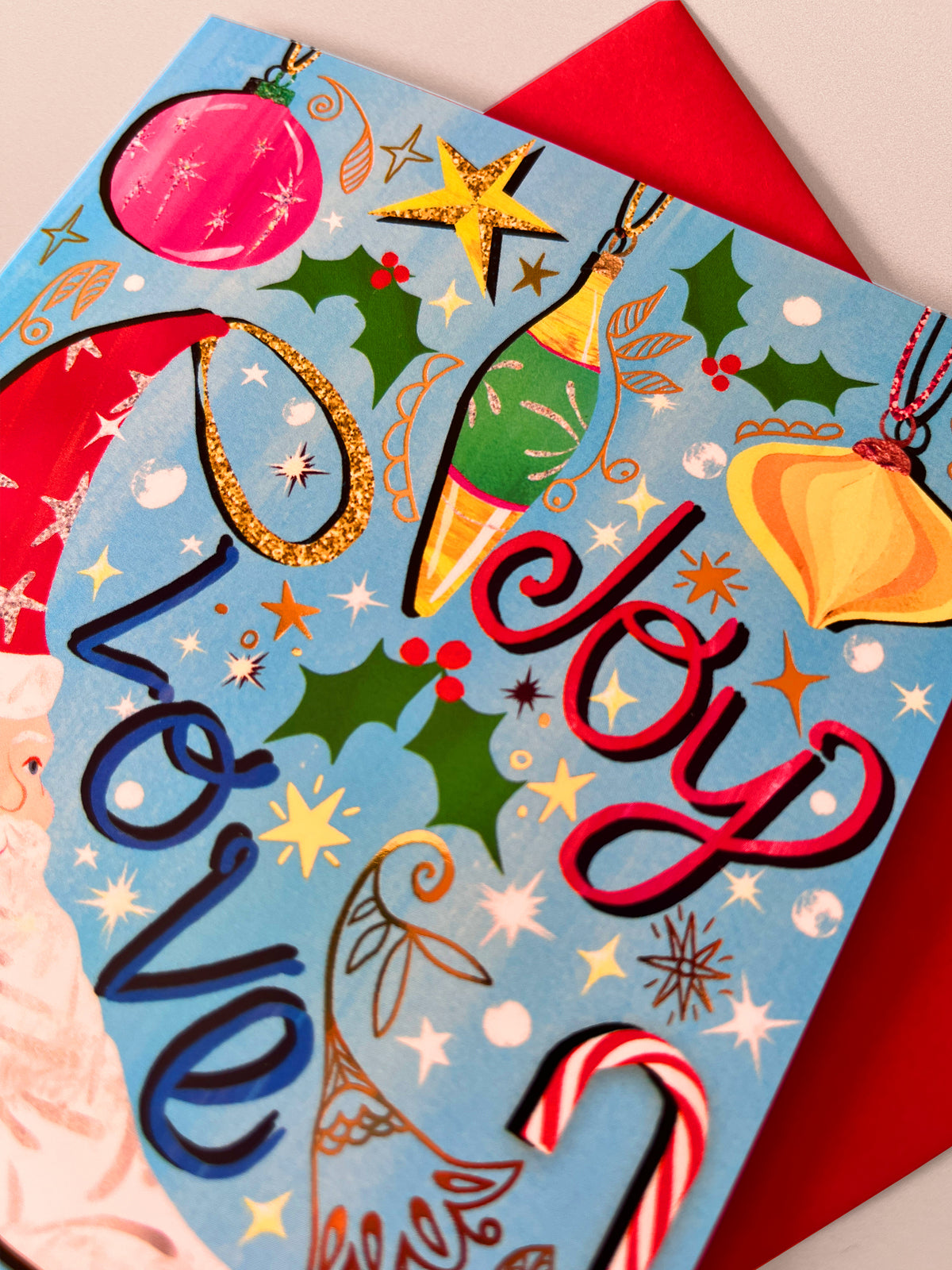 A close-up of a Christmas greeting card featuring the words 'Love' and 'Joy' in stylized lettering, decorated with festive ornaments, holly, stars, candy canes, and a red envelope behind it.