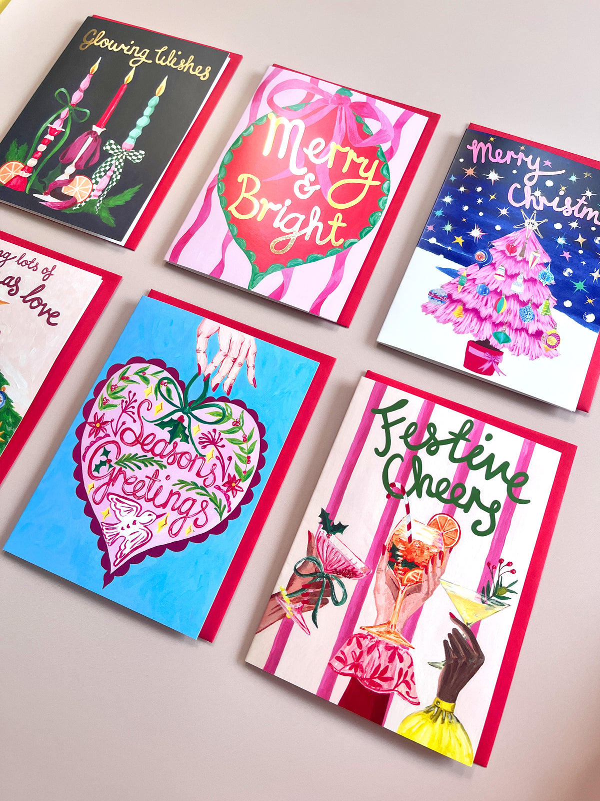 Six colorful Christmas greeting cards arranged on a light beige surface. The cards feature festive illustrations including candles, ornaments, a decorated Christmas tree, a heart with 'Merry & Bright' and 'Seasons Greetings', and cocktail glasses with 'Festive Cheers'.