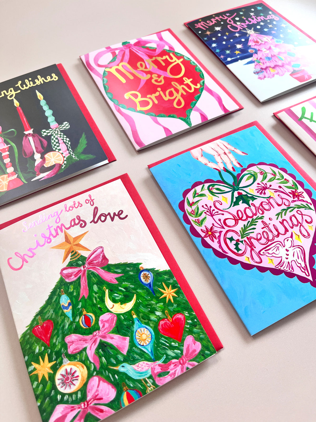 Six Christmas greeting cards arranged on a light beige surface, each featuring colorful holiday illustrations such as decorated trees, hearts, stars, and festive text.