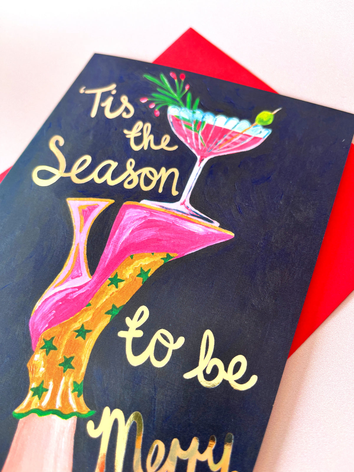A close-up of a dark navy greeting card with gold script text reading 'Tis the Season to be Merry' and a colorful illustration of a pink high heel and a pink cocktail glass, layered over a bright red envelope on a light background.