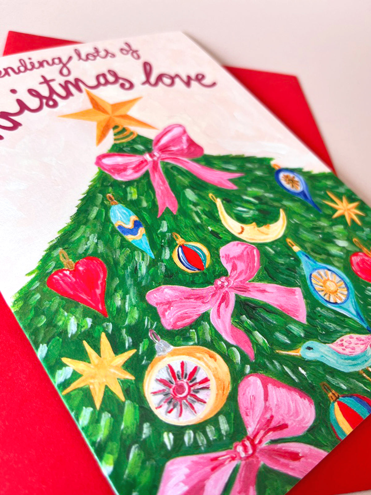 A close-up of a Christmas greeting card featuring a hand-painted green Christmas tree with pink bows, colorful ornaments, and a gold star, layered over a red envelope. The top of the card reads 'sending lots of Christmas love'.