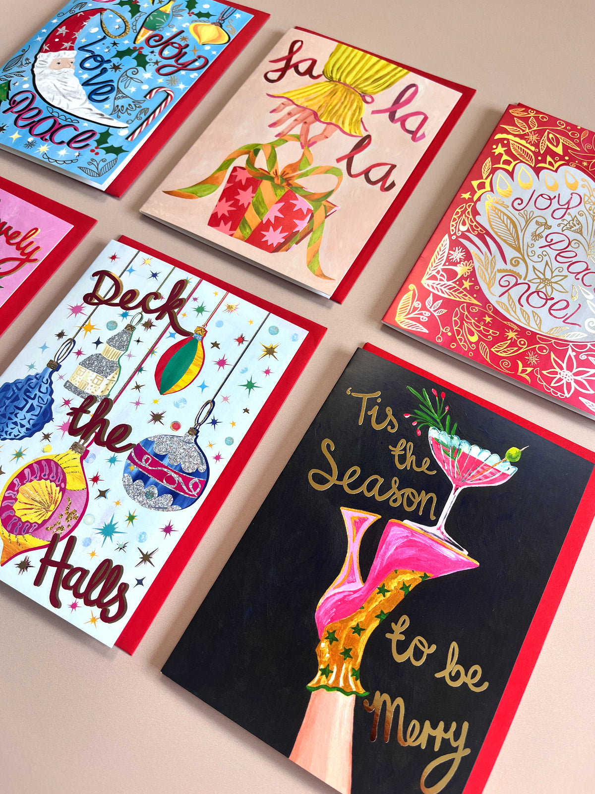 Six Christmas-themed greeting cards arranged in a flat-lay pattern on a light beige background. The cards feature colorful festive illustrations, including Santa, gifts, ornaments, a cocktail glass, and holiday phrases like 'Fa la la la', 'Deck the Halls', and 'Tis the Season to be Merry'.