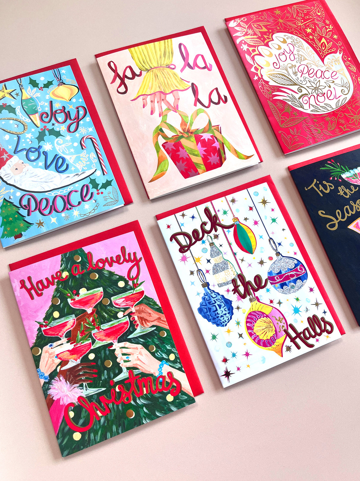 Six Christmas greeting cards arranged in a grid on a pale pink background. The cards feature festive illustrations and holiday greetings including 'Joy Peace & Love,' 'Fa la la la,' 'Joyful Peace Here,' 'Have a lovely Christmas,' 'Deck the Halls,' and 'Tis the Season for.