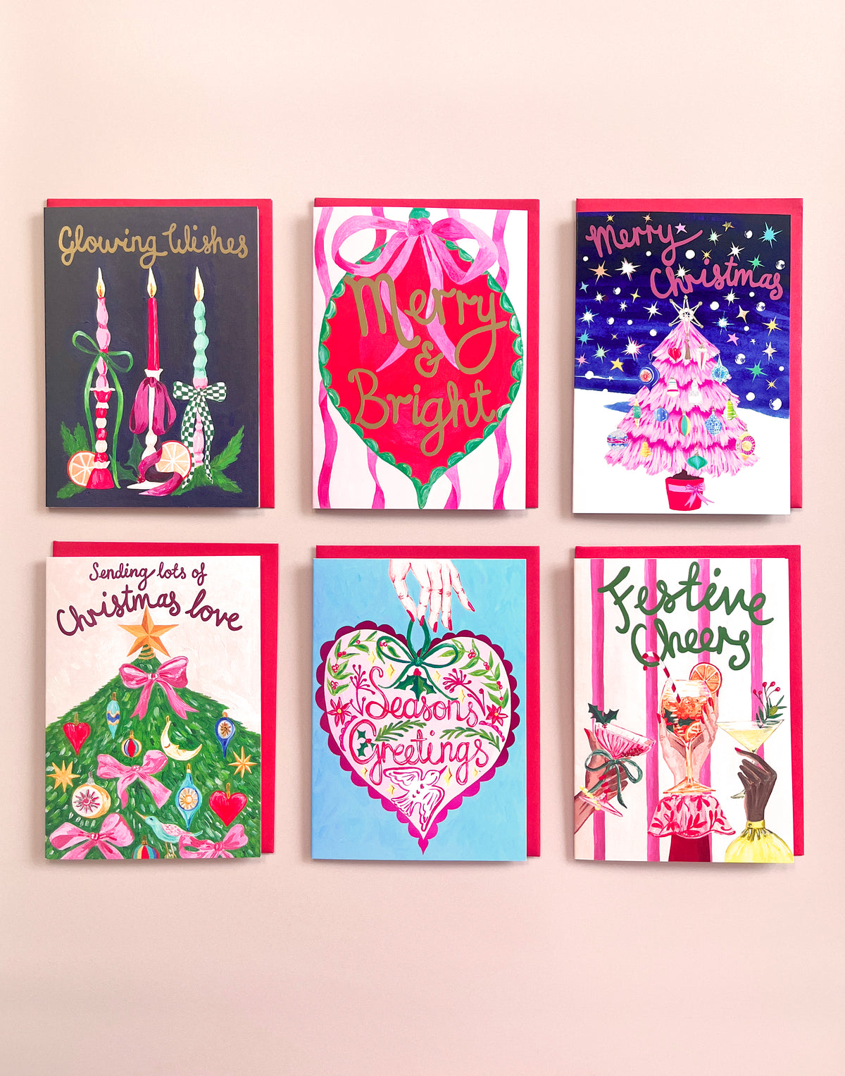 Six colorful Christmas greeting cards arranged in a 2x3 grid on a light pink wall. The cards feature festive illustrations and holiday greetings including candles, ornaments, a decorated tree, and seasonal phrases.
