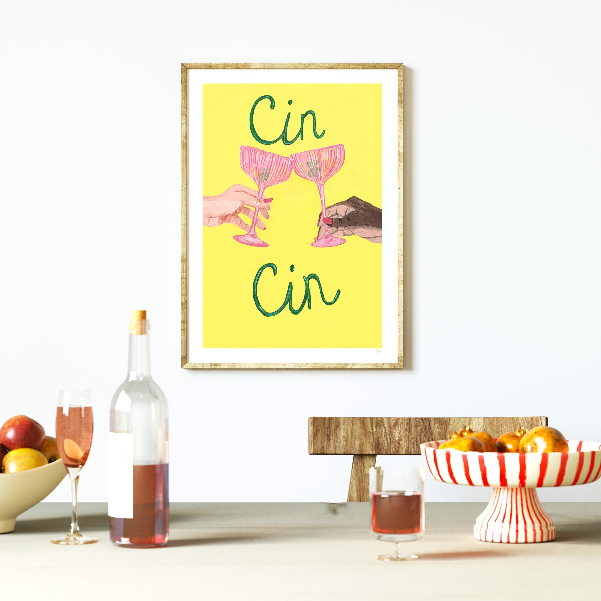 Framed artwork with two hands clinking pink glasses against a yellow background, displayed on a table with wine and fruit.