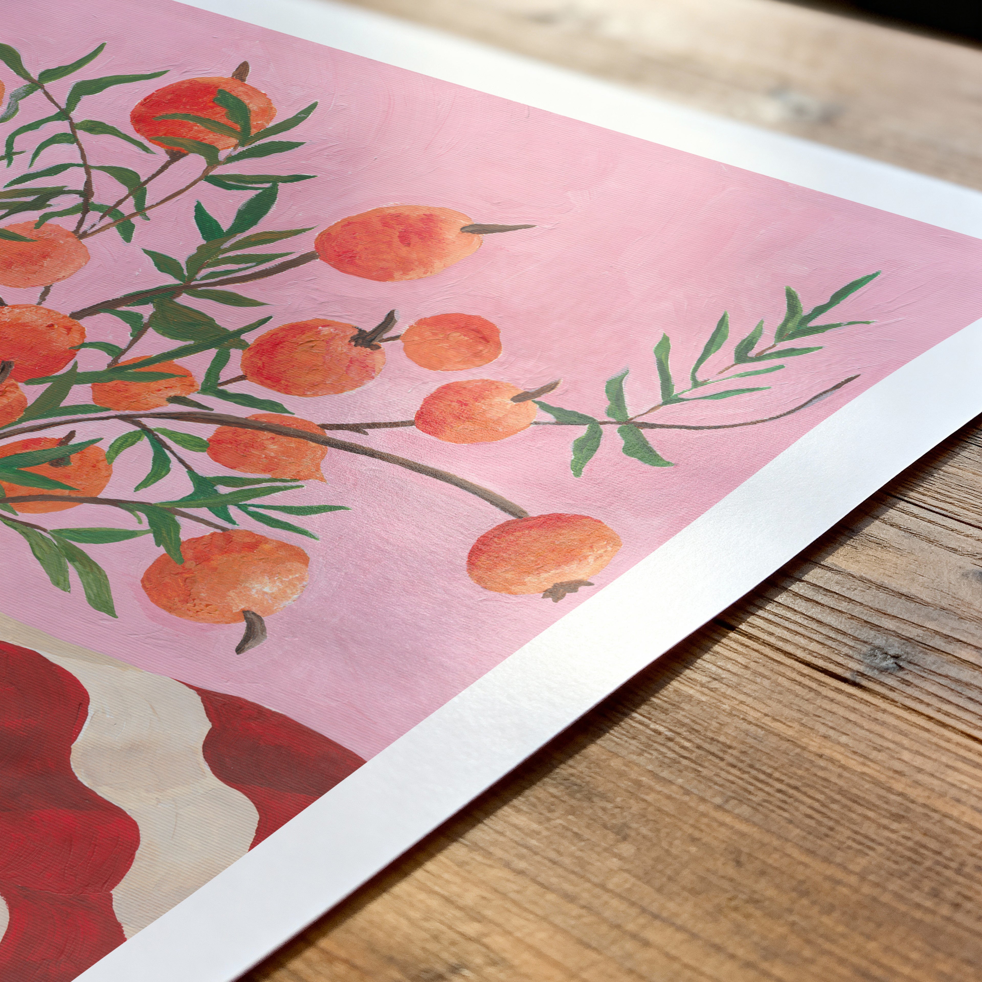 Art print of peaches on a branch with green leaves on a pink background, placed on a wooden surface.