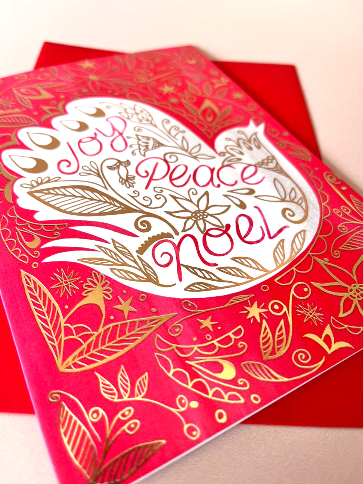 A red Christmas greeting card with gold foil floral details and a white heart shape containing the words 'Joy, Peace, and Love' in red script, shown with a matching red envelope on a light beige background.