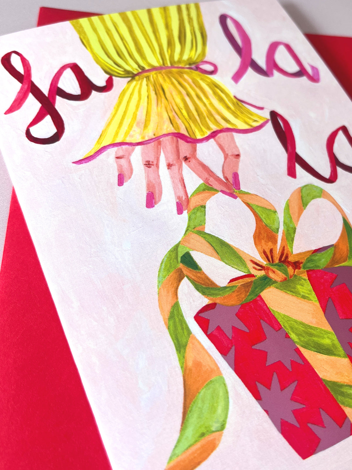 A close-up of a printed illustration on a white card or envelope, showing a stylized hand with pink nails and a yellow pleated sleeve, holding a red gift box with green and orange ribbon. Red script lettering appears across the top, and a red background is visible around the card.