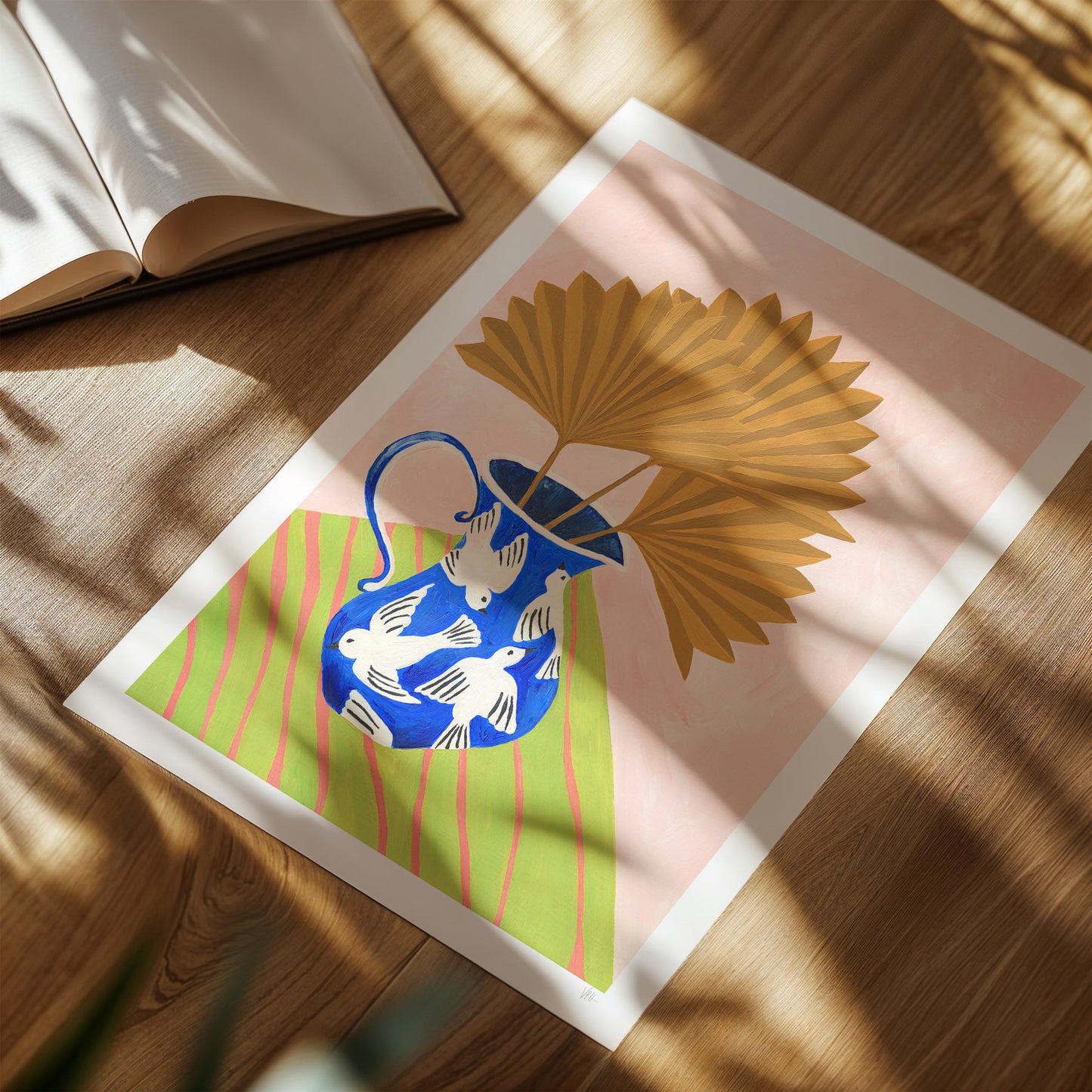 Framed artwork with a blue vase and white birds on a pink background, placed on a wooden surface.
