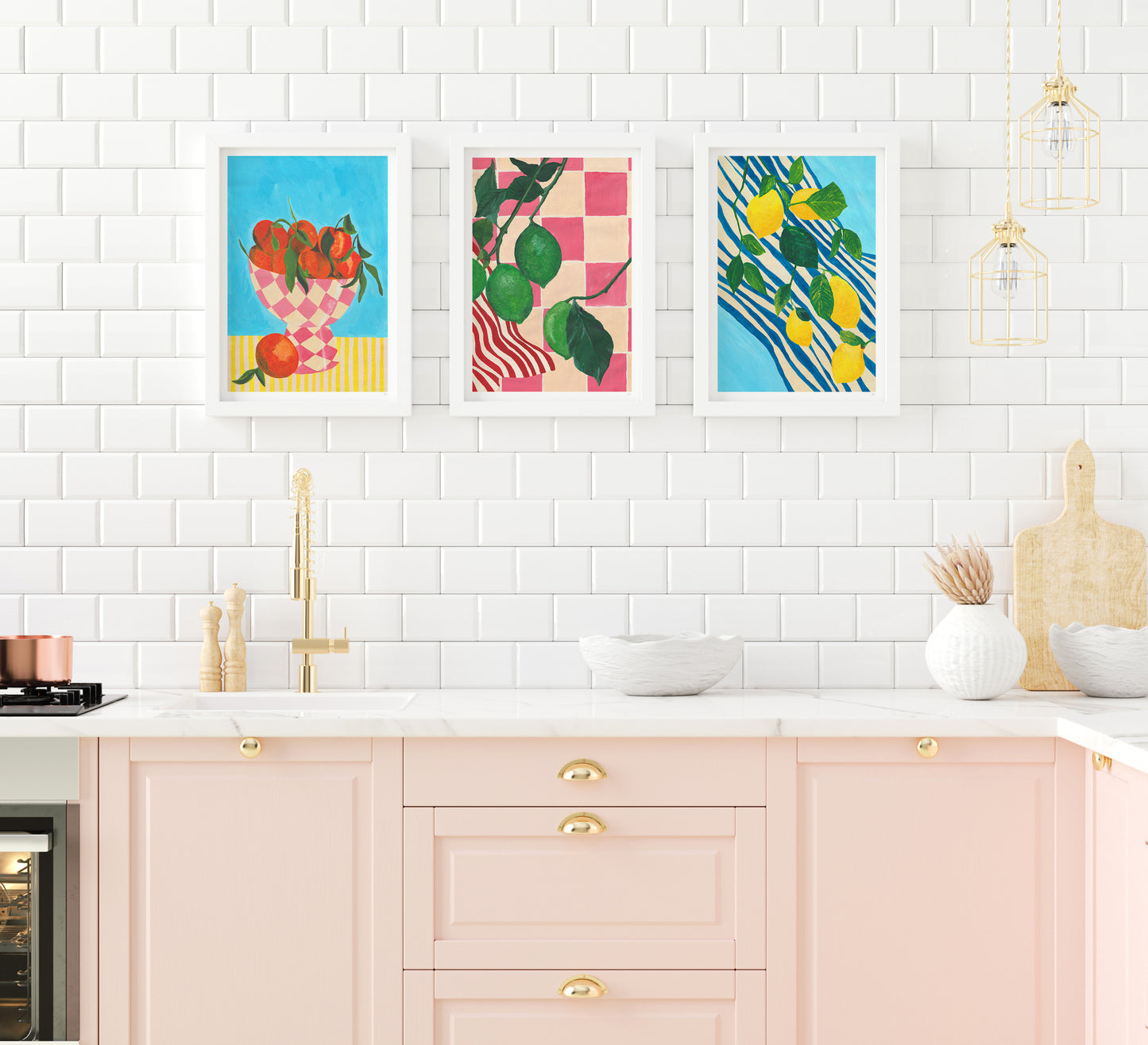 A briight white and soft pink kitchen with gold hardware displaying a set of 3 colourful citrus fruit prints above the sink