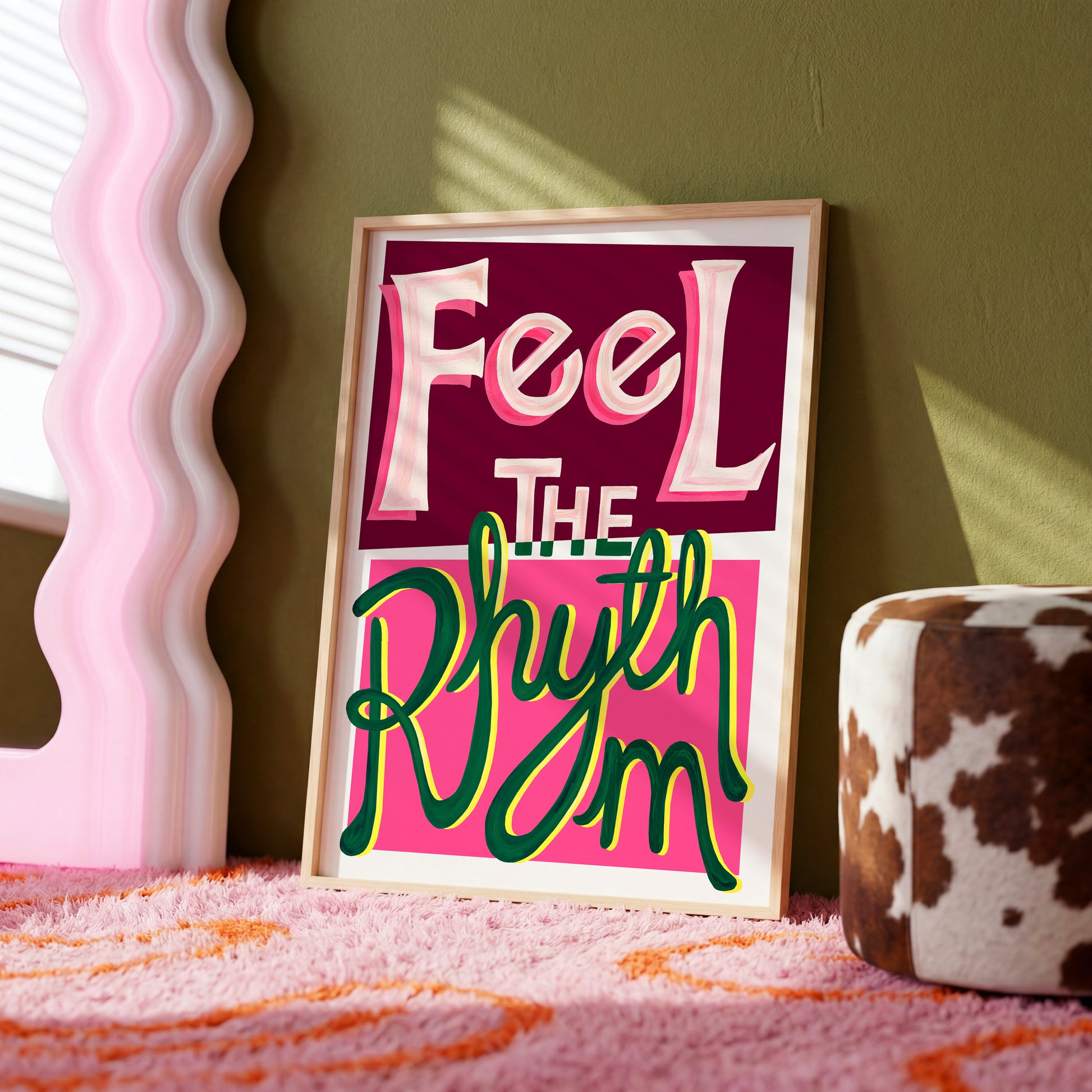 Framed artwork with 'Feel The Rhythm' text on a pink background, placed on a surface with a decorative mirror and cow print item in the background.