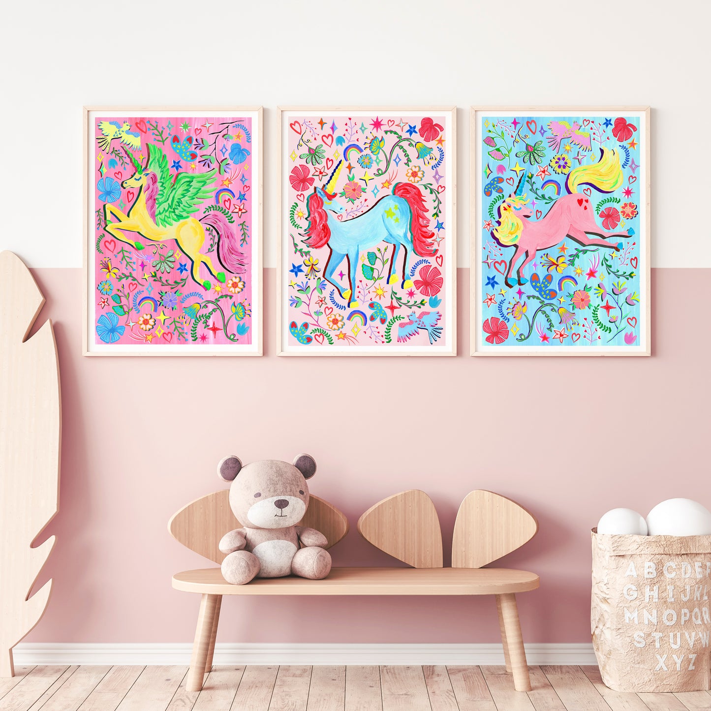 Three colorful unicorn prints on a pink wall above a wooden bench with a teddy bear.