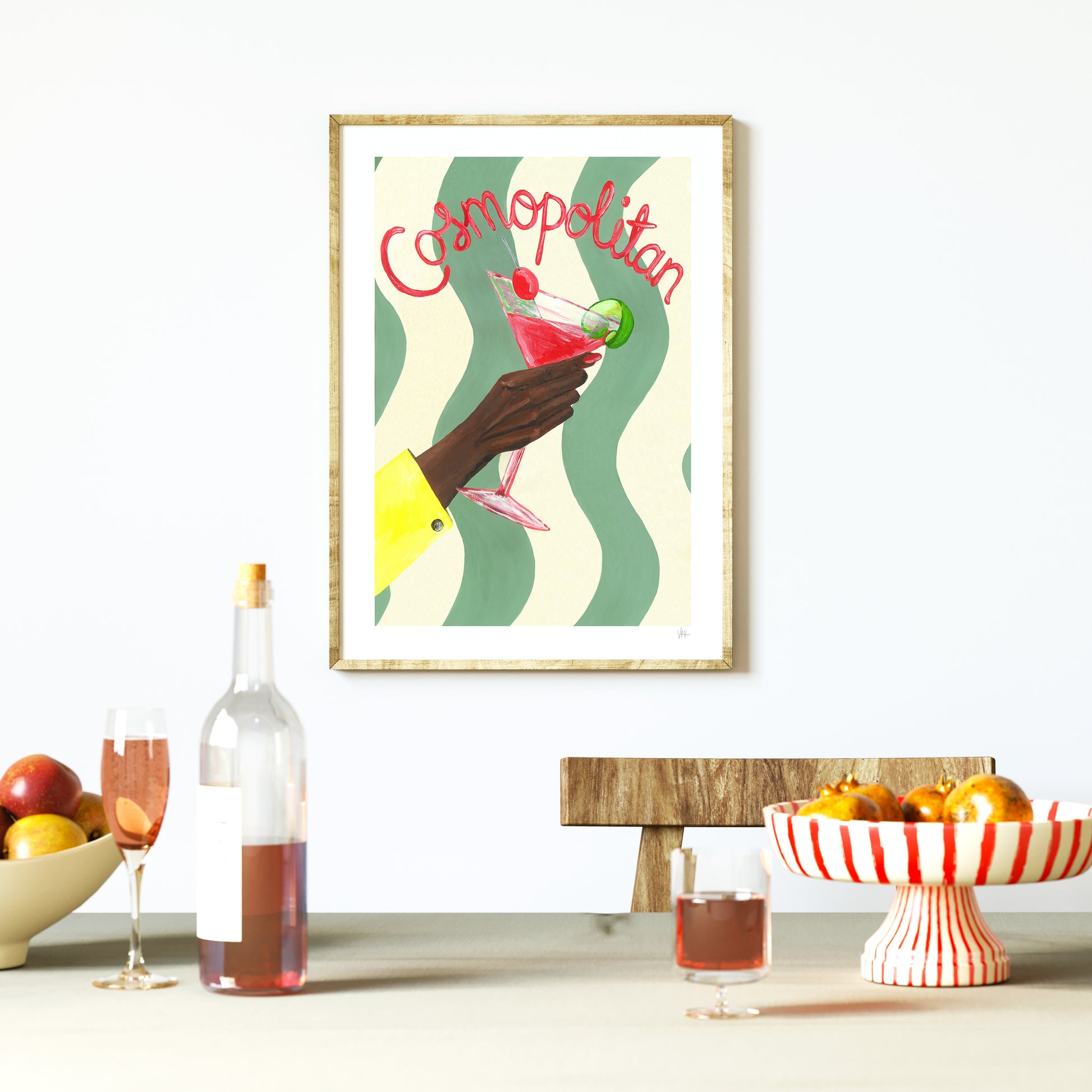 Framed artwork of a hand holding a Cosmopolitan cocktail with a bottle and fruit bowl on a table.
