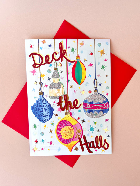 A square Christmas greeting card with the phrase 'Deck the Halls' in red script, decorated with colorful hanging ornaments and star motifs on a white background, styled on top of a red envelope against a soft pink backdrop.