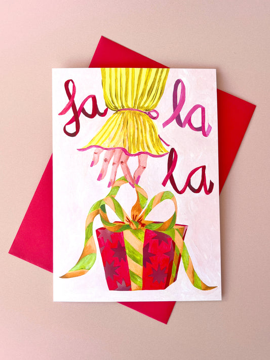 A festive greeting card featuring the words 'la la la' in pink and red script above an illustration of a yellow pleated dress and a red gift box with green and gold ribbon, displayed on top of a red envelope against a beige background.