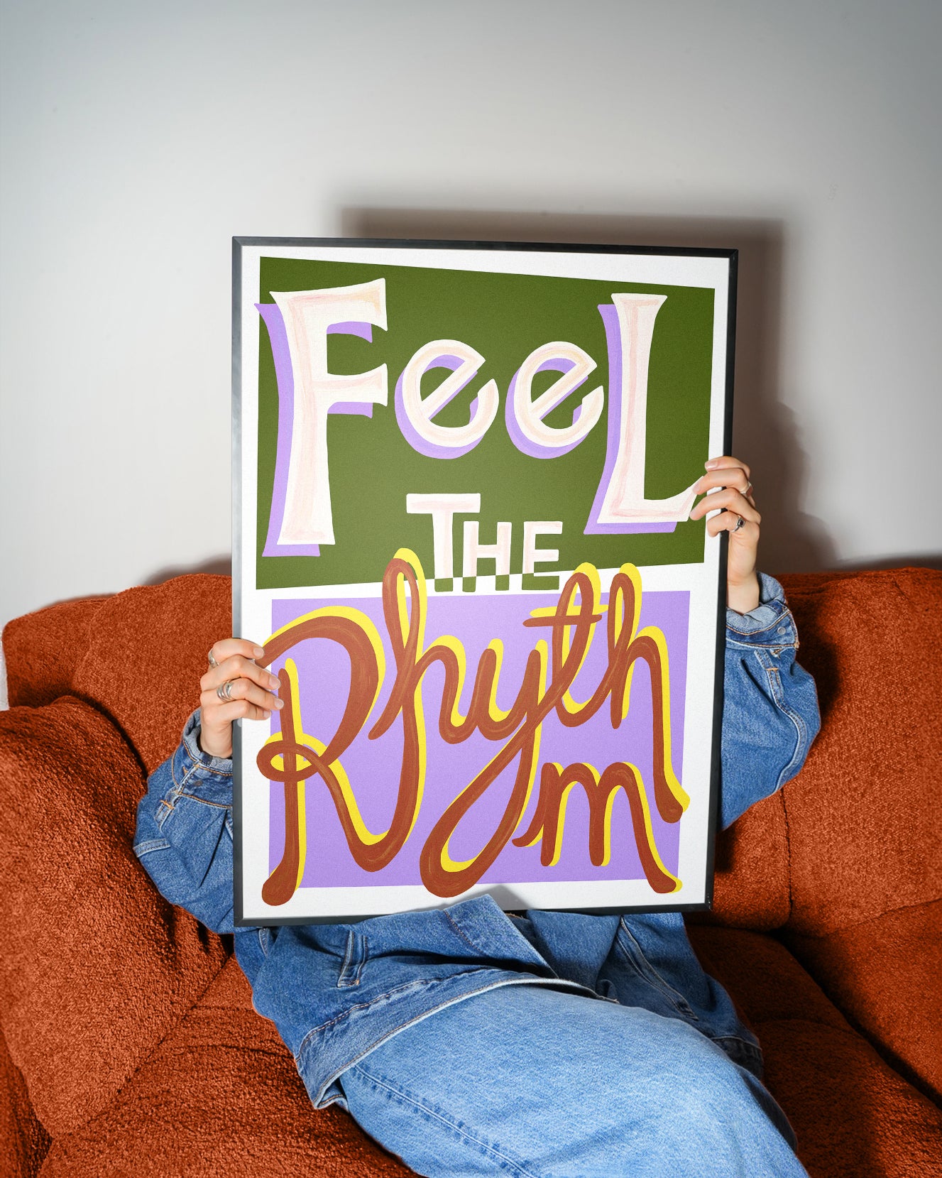 Person holding a framed music typography poster with 'Feel The Rhythm' text on a couch.